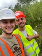 Uśmiechnięci pracownicy w kaskach i kamizelkach odblaskowych na tle zieleni. Selfie z budowy, widoczne logo firmy na kasku.