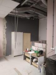 Wnętrze w trakcie remontu: szara ściana, częściowo wykończona ściana z widocznym szpachlowaniem, szafki i materiały budowlane. Widoczne instalacje pod sufitem.