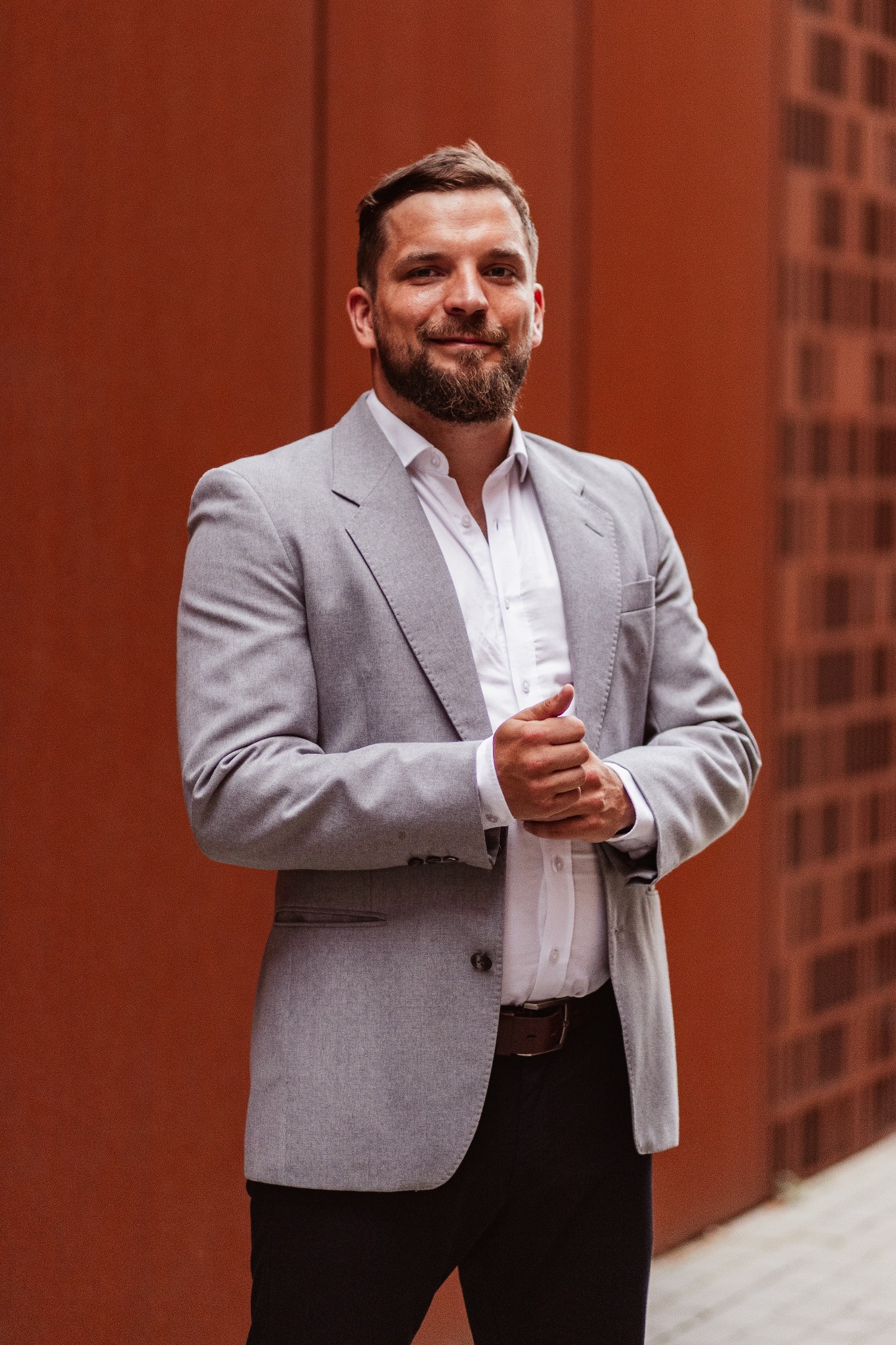 Uśmiechnięty mężczyzna z brodą w szarej marynarce i białej koszuli, stojący na tle ceglanej ściany. Profesjonalny wizerunek doradcy finansowego.