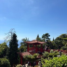 Minda Dorota - Egzotyczny ogród z czerwoną pagodą i turystami na tle błękitnego nieba i oceanu. Bujna roślinność, palmy i drzewa tworzą tropikalny klimat.