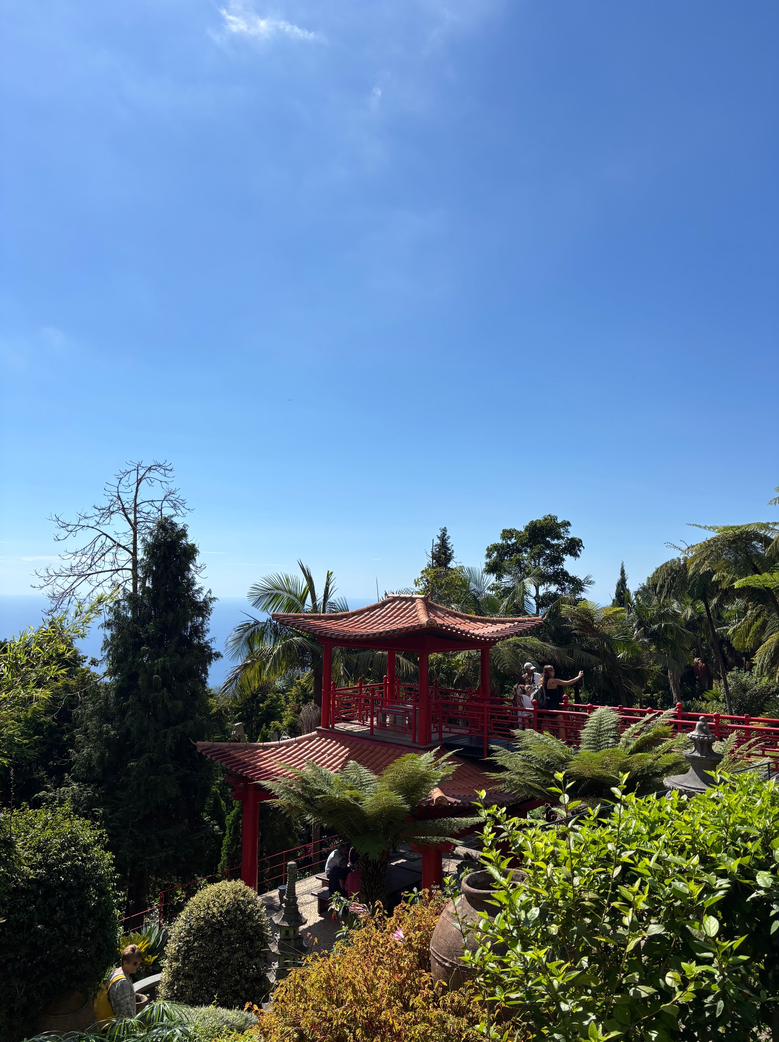 Egzotyczny ogród z czerwoną pagodą i turystami na tle błękitnego nieba i oceanu. Bujna roślinność, palmy i drzewa tworzą tropikalny klimat.