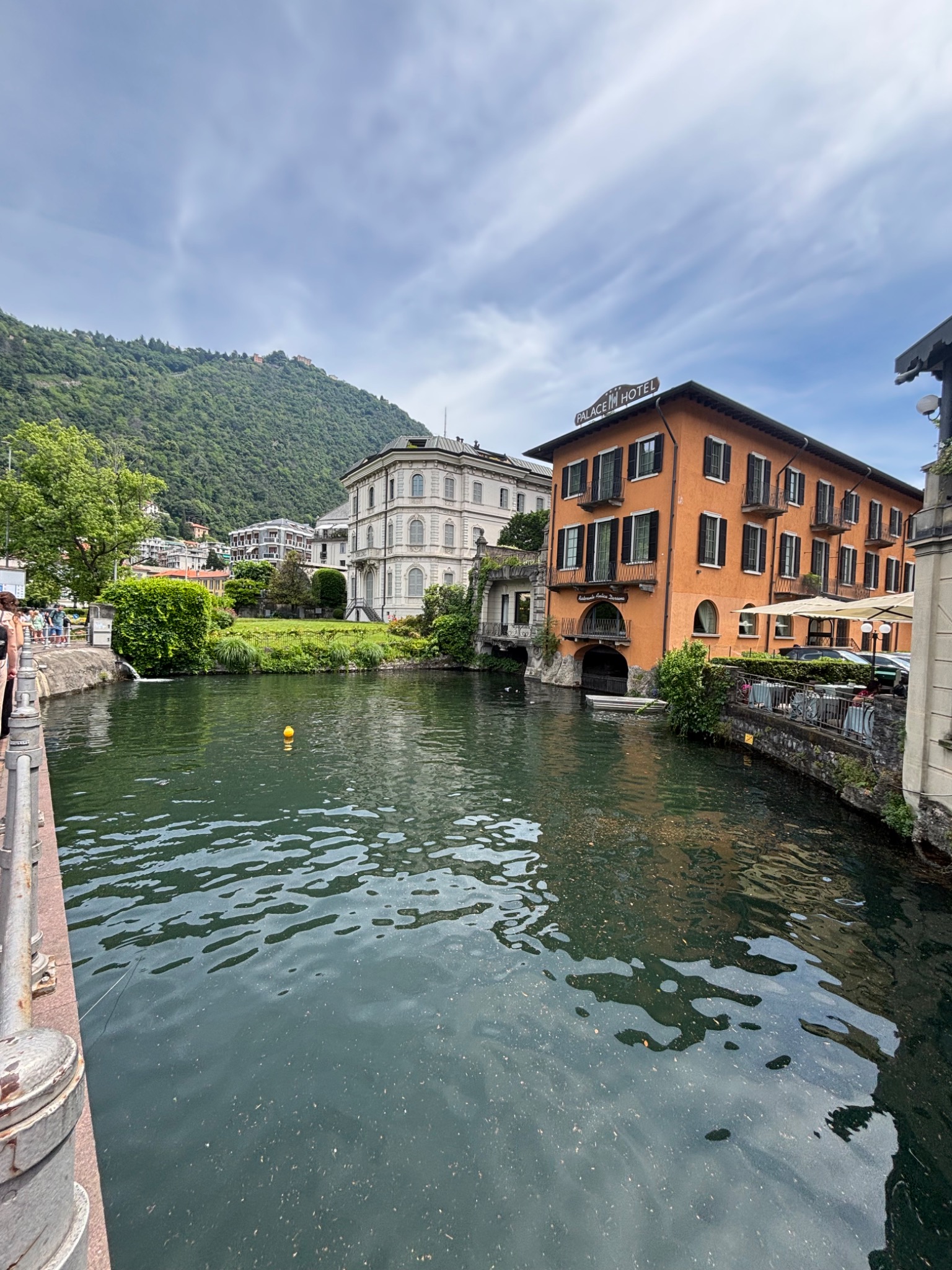 Widok na jezioro z budynkami w tle, w tym pomarańczowy hotel z napisem 'PALACE HOTEL' na dachu. Woda odbija niebo i budynki, tworząc malowniczy krajobraz.