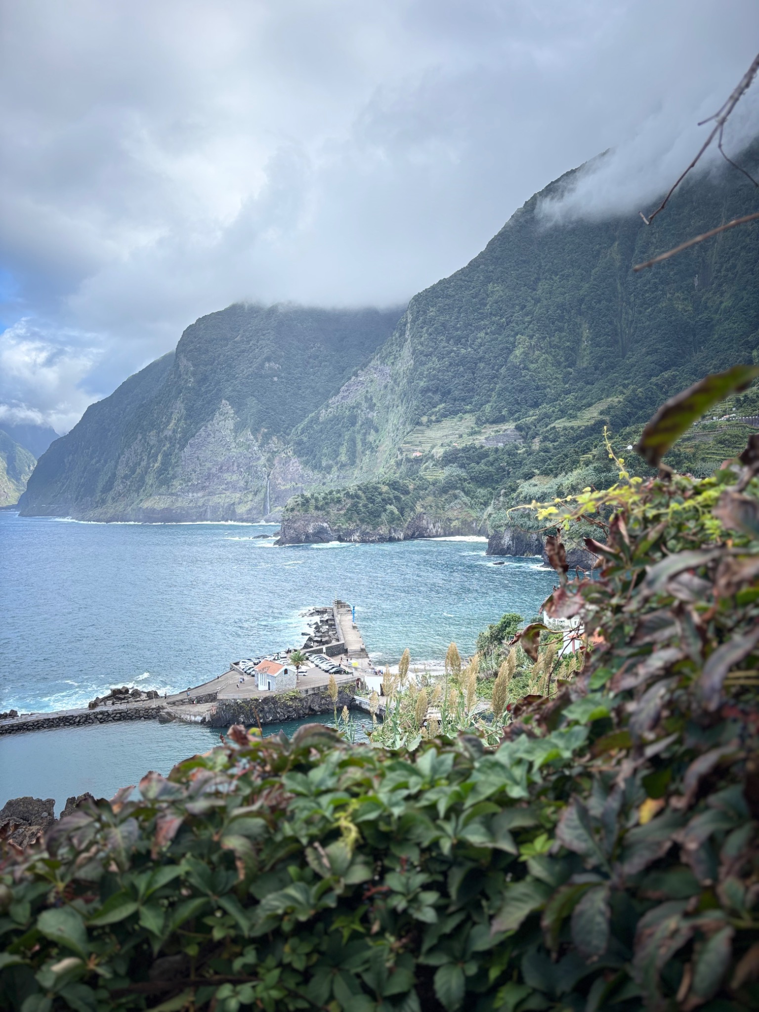Malownicza panorama: zielone góry schodzą do błękitnego oceanu, z widocznym portem i roślinnością na pierwszym planie. Mgliste chmury otulają szczyty.