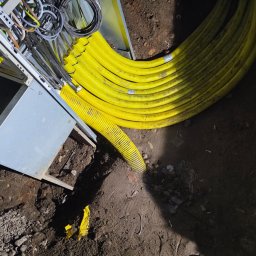 V.S.E.G. Sp. z o.o. - Żółte rury karbowane prowadzone w ziemi obok metalowej skrzynki z przewodami. Widoczne okablowanie i elementy instalacji w ziemnym wykopie.