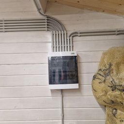 V.S.E.G. SPÓŁKA Z OGRANICZONĄ ODPOWIEDZIALNOŚCIĄ - Rozdzielnia elektryczna natynkowa z szarymi przewodami w peszlach, zamontowana na ścianie z boazerii pomalowanej na biało. Widoczne zabezpieczenia i fragment worka jutowego.