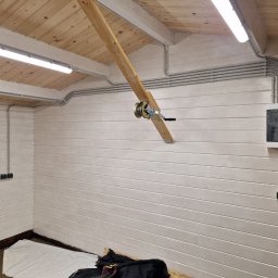V.S.E.G. SPÓŁKA Z OGRANICZONĄ ODPOWIEDZIALNOŚCIĄ - Szara instalacja elektryczna natynkowa w garażu z drewnianym sufitem i białą boazerią, widoczna skrzynka rozdzielcza i oświetlenie LED.