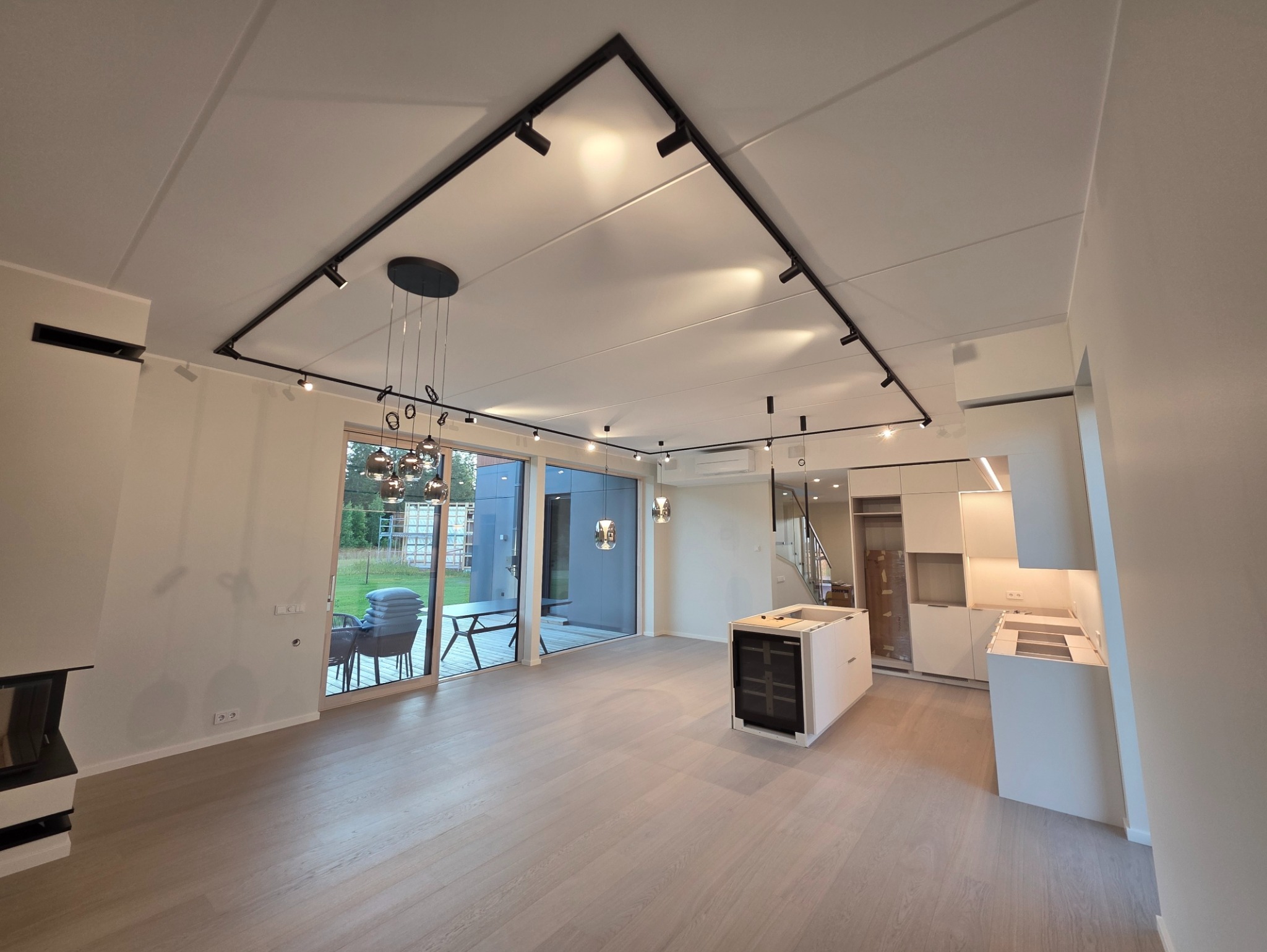 Nowoczesny salon z oświetleniem szynowym w kształcie trójkąta, wiszącą lampą nad stołem i minimalistyczną zabudową kuchenną w odcieniach bieli i szarości.