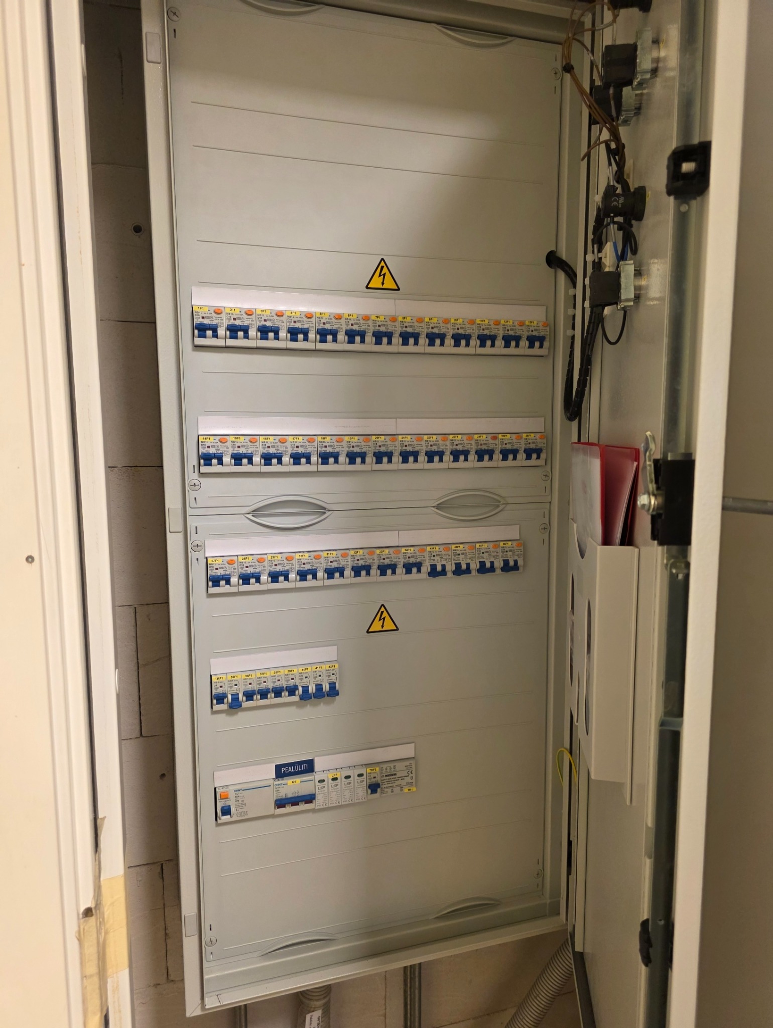 Otwarta skrzynka elektryczna z bezpiecznikami, widoczne okablowanie i elementy montażowe. Skrzynka zamontowana na ścianie z surowego betonu komórkowego.