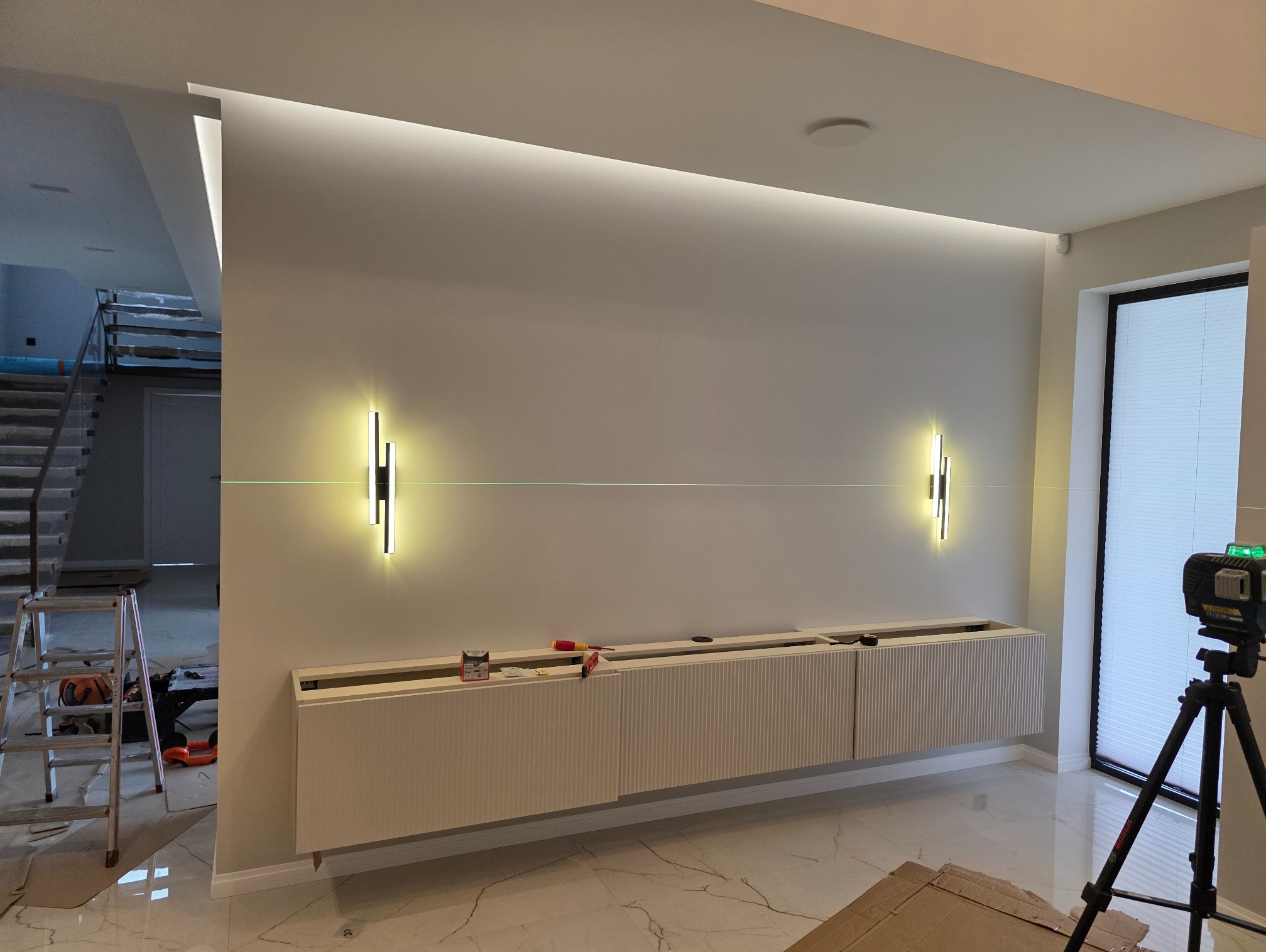 Montaż oświetlenia ściennego nad szafką RTV w nowoczesnym salonie. Widoczny laser krzyżowy, drabina i narzędzia. Minimalistyczny design, ciepłe światło.