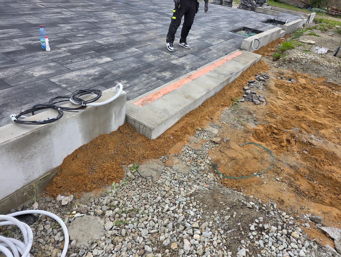 Kable w peszlu ułożone wzdłuż betonowego obrzeża na posesji z kostki. Widoczne przygotowania do montażu, ziemia i kamienie. Prace ziemne.