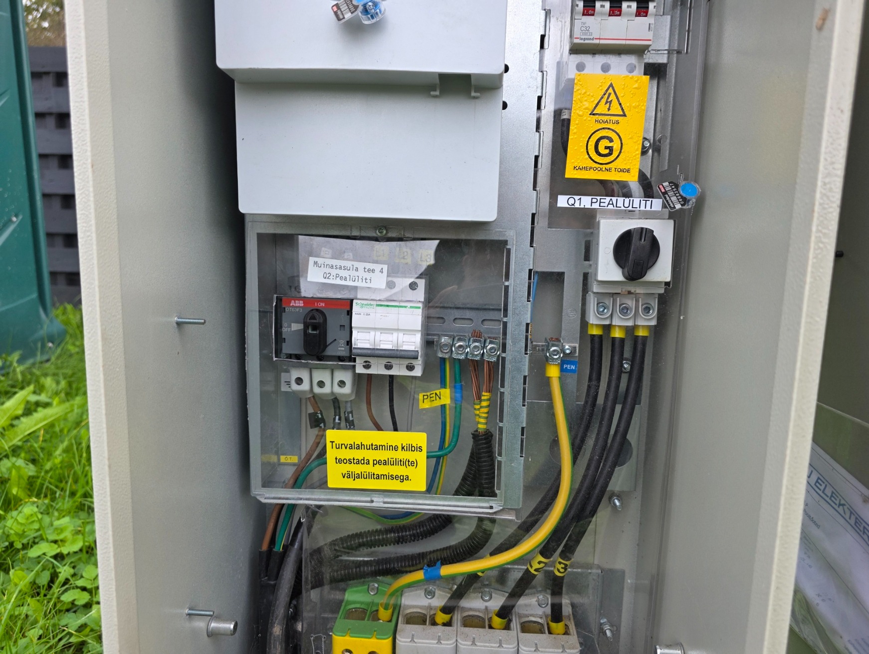 Otwarta skrzynka elektryczna z widocznymi bezpiecznikami, przewodami oznaczonymi kolorami i żółtą naklejką ostrzegawczą. Widoczne elementy instalacji elektrycznej.