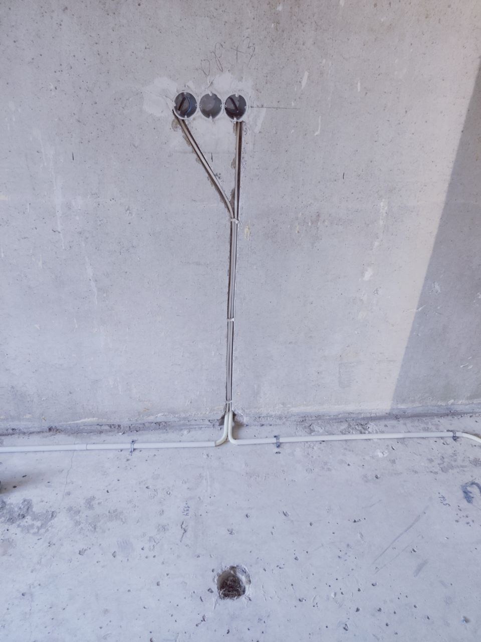 Surowa ściana z trzema otworami i wystającymi przewodami elektrycznymi, pionowo poprowadzonymi w dół. Dodatkowo widoczna pozioma rurka instalacyjna.