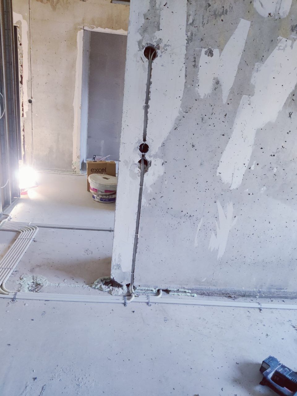 Surowy beton, pionowy kanał z kablami w ścianie. Białe rury na podłodze, puszki z zaprawą w tle. Instalacja elektryczna w trakcie prac wykończeniowych.