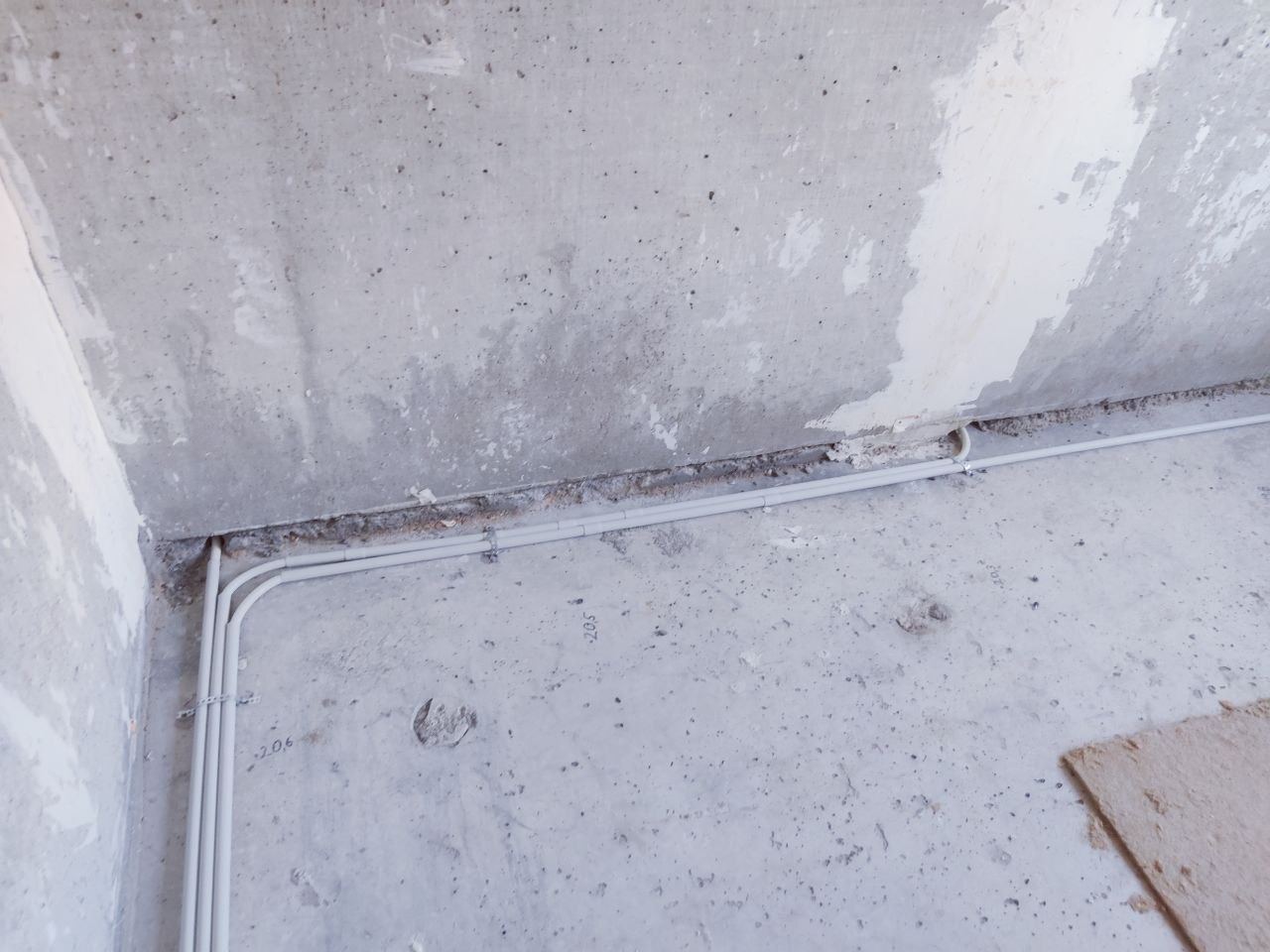 Białe rury instalacji elektrycznej prowadzone wzdłuż surowej, szarej ściany w trakcie remontu. Widoczne mocowania rur i niedoskonałości powierzchni.