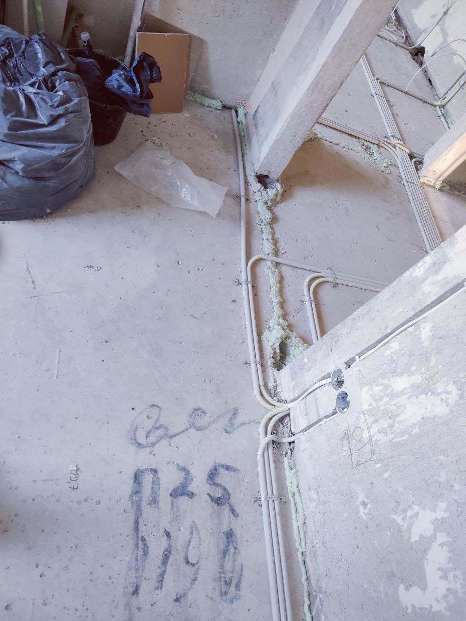 Białe rury elektryczne prowadzone wzdłuż surowej ściany, zabezpieczone pianką montażową. Na podłodze widoczne napisy wykonane ołówkiem. Surowy stan wykończenia.