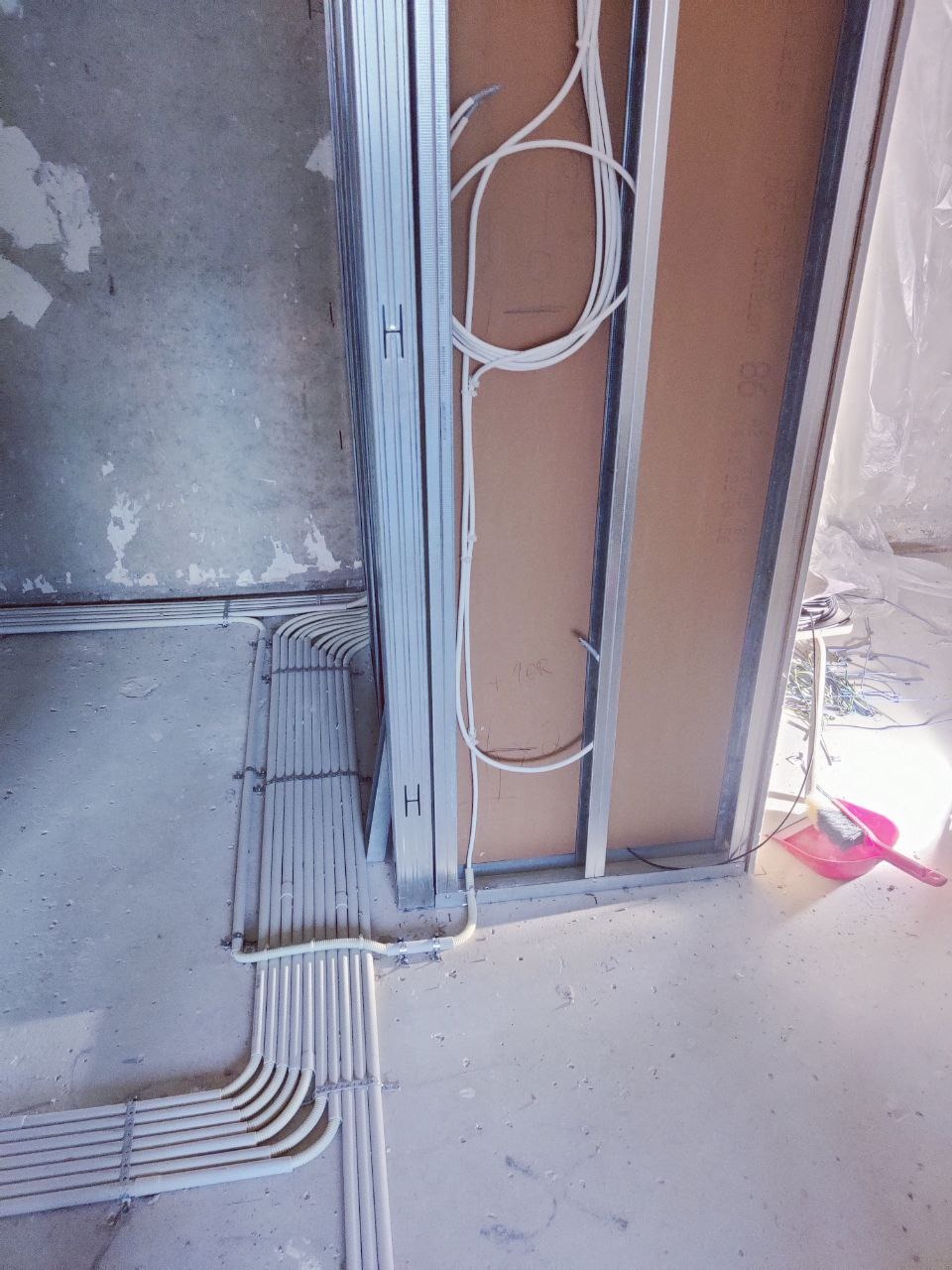 Białe rury instalacji elektrycznej ułożone wzdłuż ściany z metalowym profilem i płytą kartonowo-gipsową w surowym wnętrzu. Widoczne kable i narzędzia.