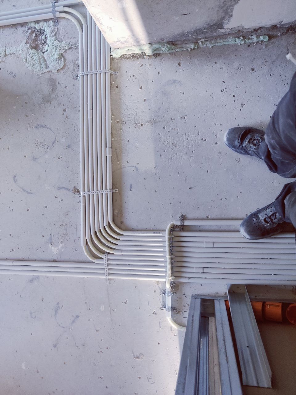 Białe rury instalacji elektrycznej ułożone równolegle na szarej, surowej ścianie. Widoczne mocowania i fragment buta osoby stojącej obok. Ujęcie z góry.