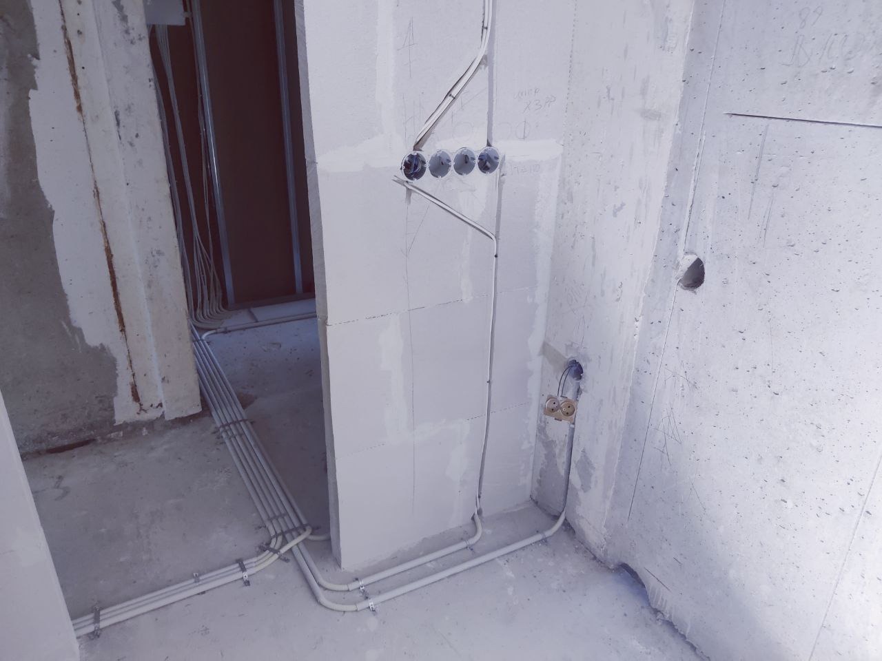 Szare rury elektryczne prowadzone po ścianie w surowym wnętrzu. Widoczne puszki instalacyjne i przewody. Ściany w stanie deweloperskim, surowy beton.