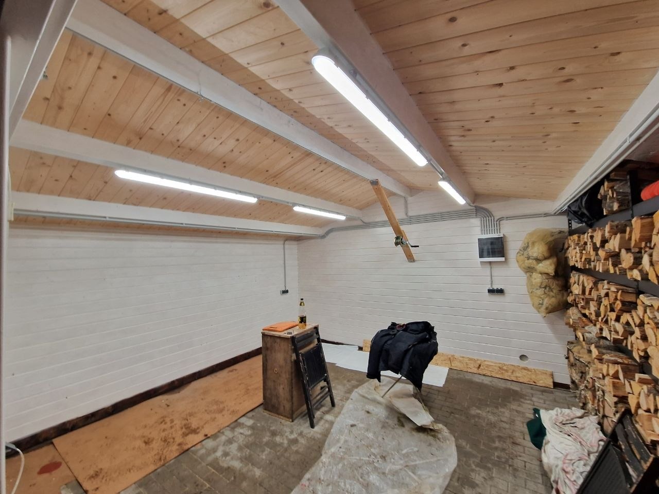 Wnętrze garażu z drewnianym sufitem i zamontowanym oświetleniem LED. Widoczne białe ściany, regał z drewnem i instalacja elektryczna.