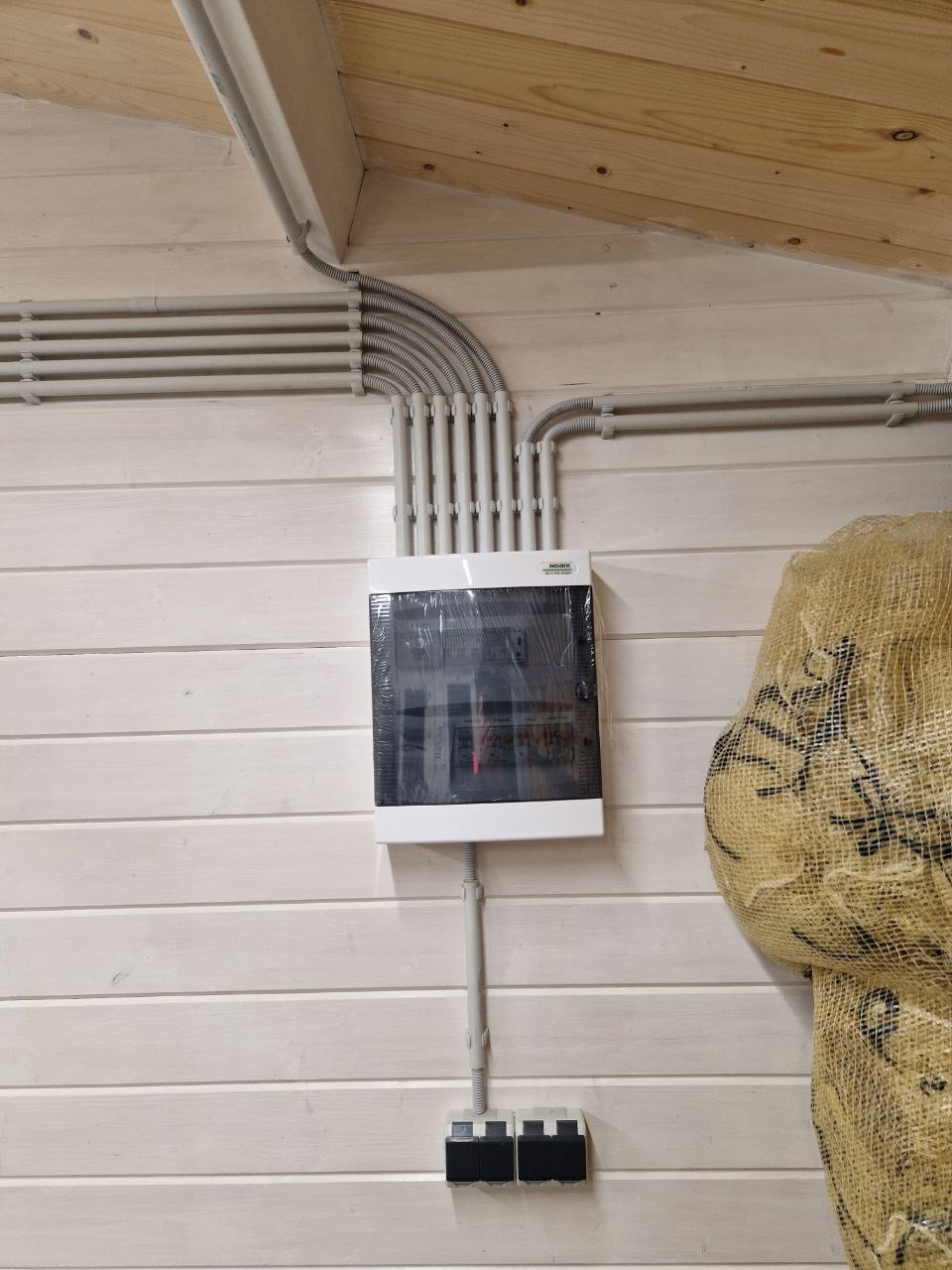 Rozdzielnia elektryczna natynkowa z szarymi przewodami w peszlach, zamontowana na ścianie z boazerii pomalowanej na biało. Widoczne zabezpieczenia i fragment worka jutowego.
