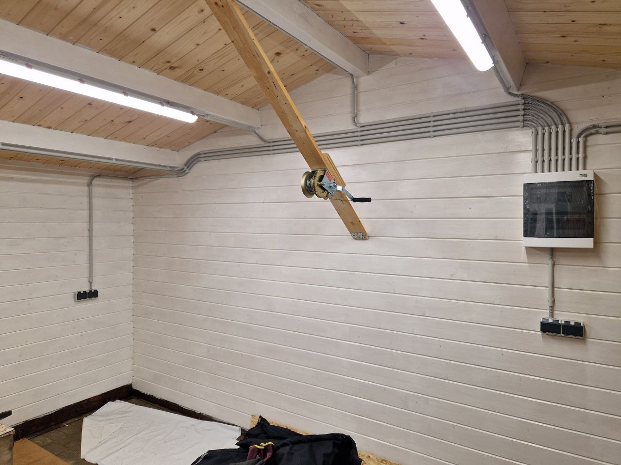 Szara instalacja elektryczna natynkowa w garażu z drewnianym sufitem i białą boazerią, widoczna skrzynka rozdzielcza i oświetlenie LED.