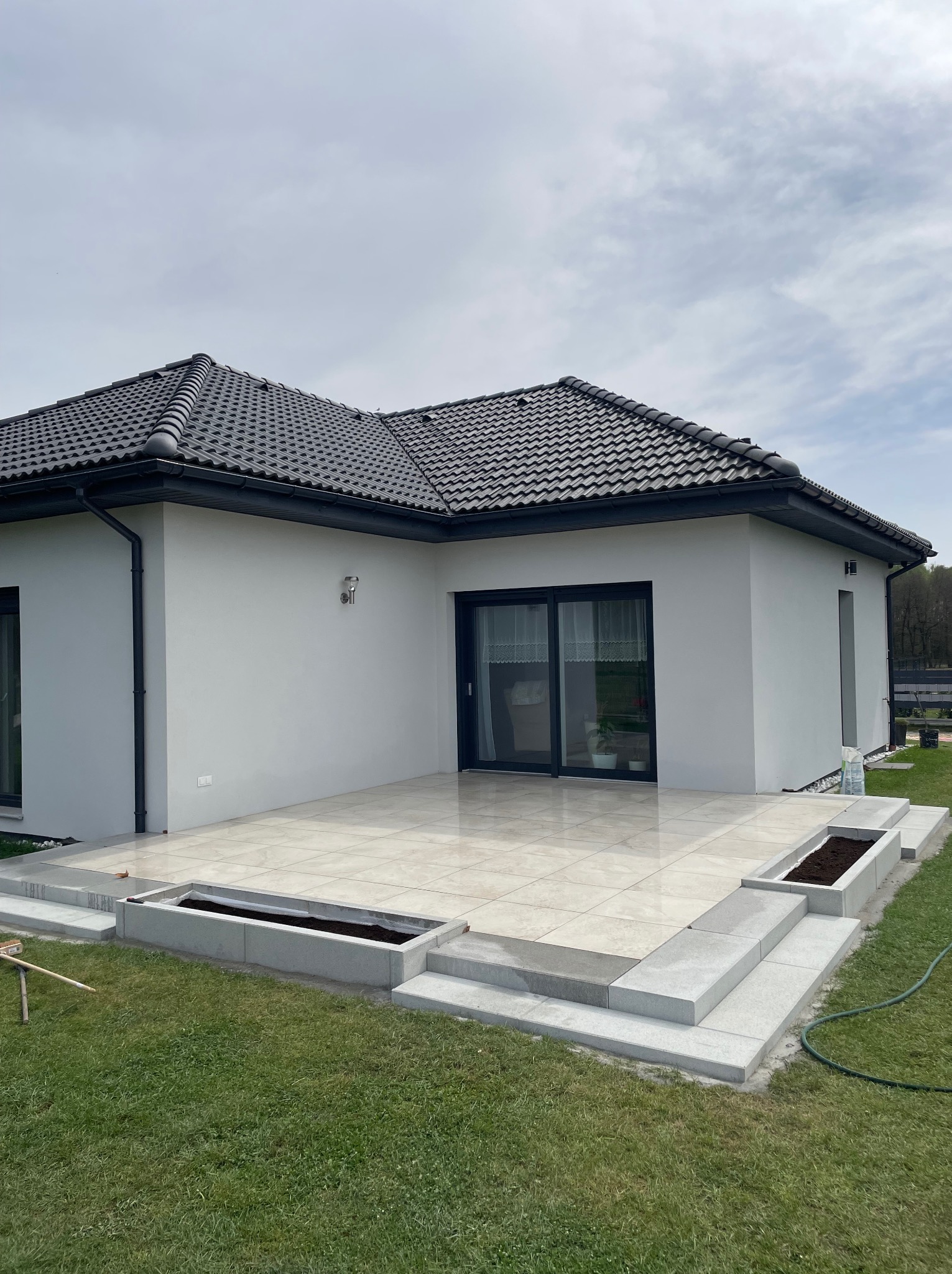 Jasny taras z płytek przy nowoczesnym domu z ciemną dachówką. Schody z szarej kostki prowadzą do trawnika. Widoczne rabaty kwiatowe.