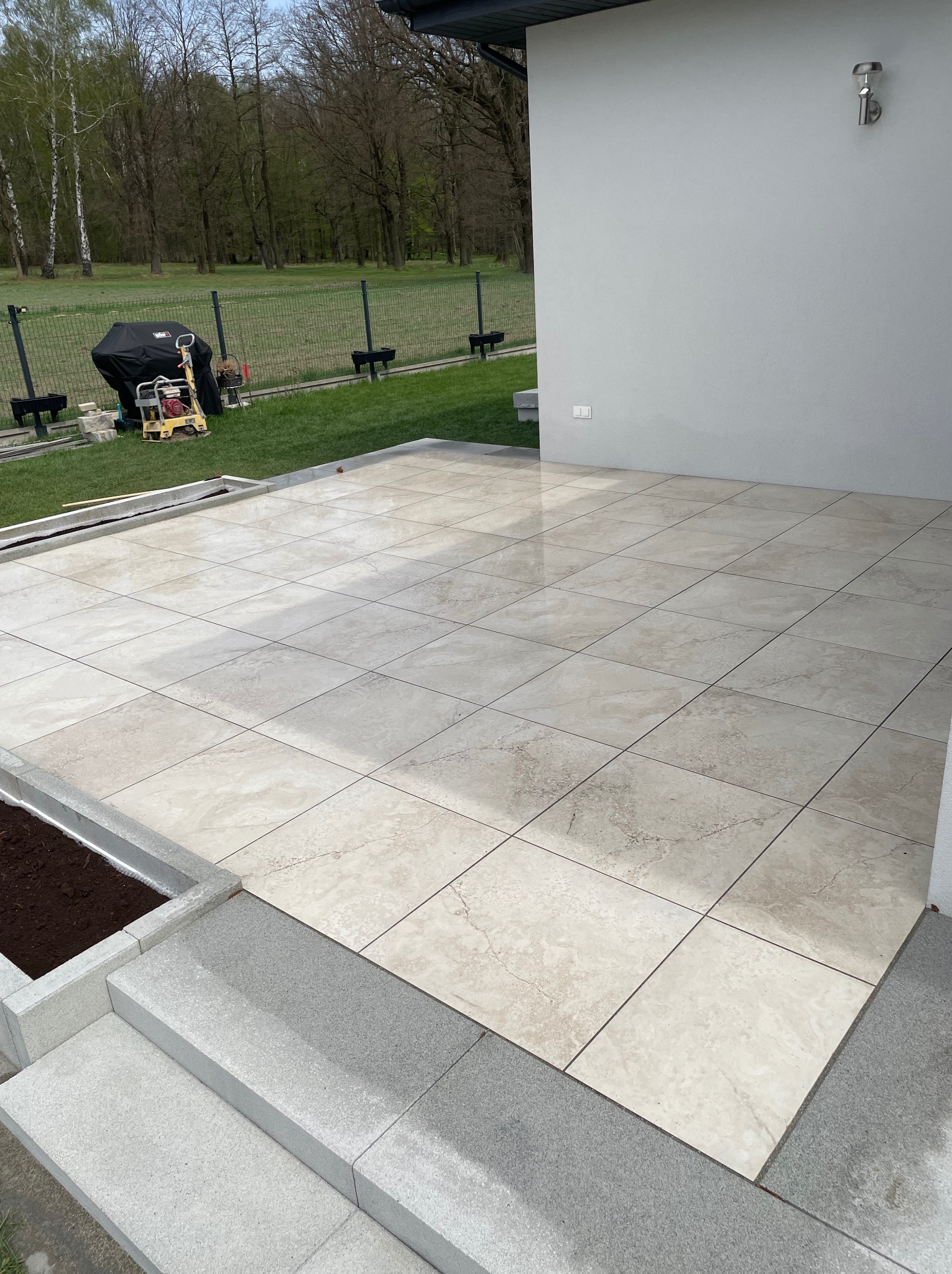Jasne, kwadratowe płytki tarasowe ułożone na zewnątrz budynku. Widoczne schody z szarego kamienia i fragment trawnika z urządzeniem budowlanym w tle.