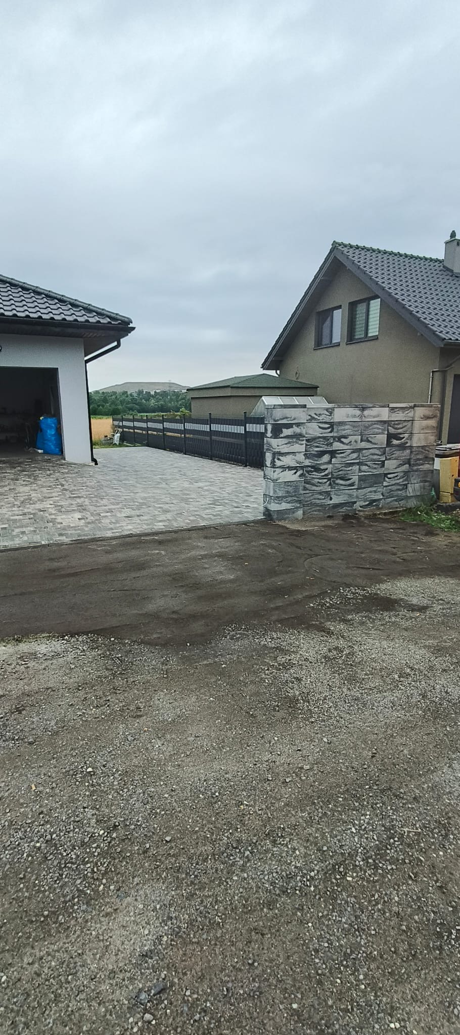 Ułożona kostka brukowa na podjeździe domu jednorodzinnego, widoczny fragment ogrodzenia i sterta kostki. Pochmurne niebo.