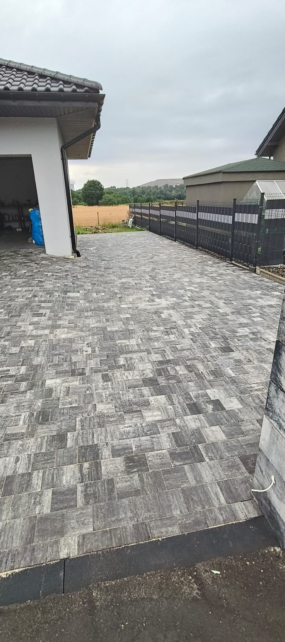 Szara kostka brukowa ułożona na podjeździe, widok z perspektywy, zarys domu i ogrodzenie w tle, pochmurne niebo.
