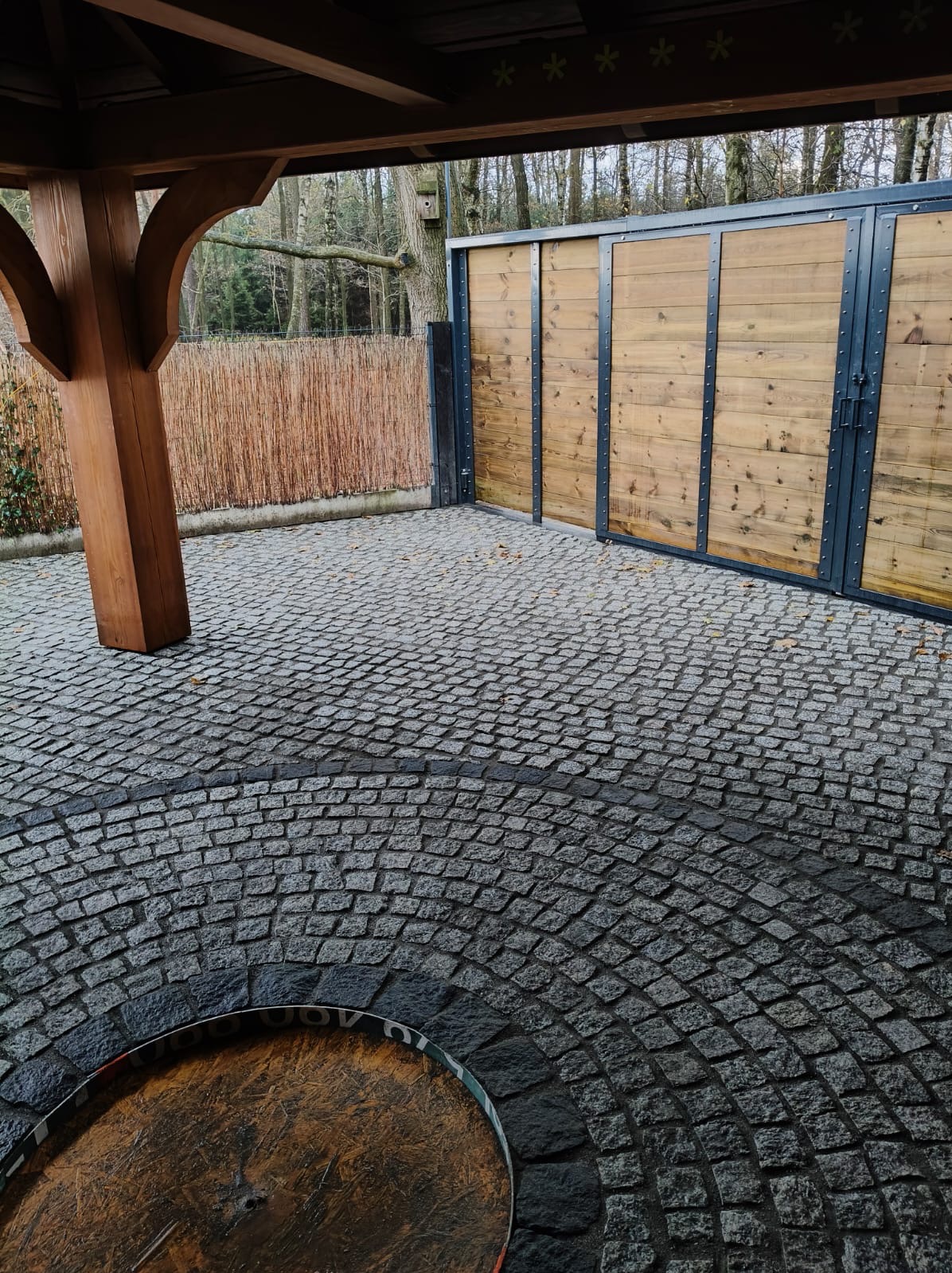 Szara kostka granitowa ułożona w okrągły wzór pod drewnianą altaną, z drewnianą bramą w tle. Widoczne detale faktury kostki i naturalne oświetlenie.