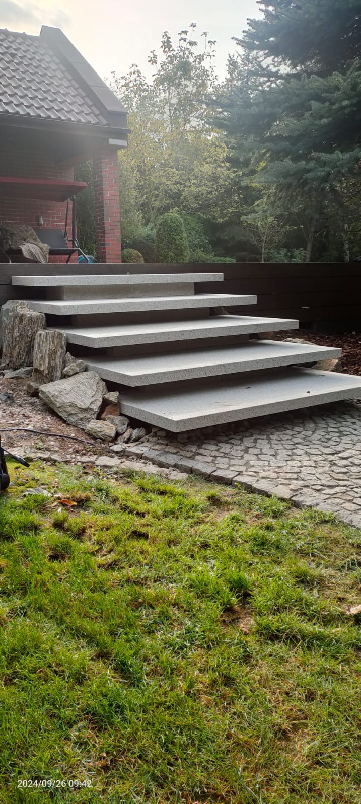 Granitowe schody zewnętrzne prowadzące do domu, otoczone kamieniami i zielenią. Widoczna kostka brukowa i drewniane ogrodzenie w tle. Ujęcie w pochmurny dzień.