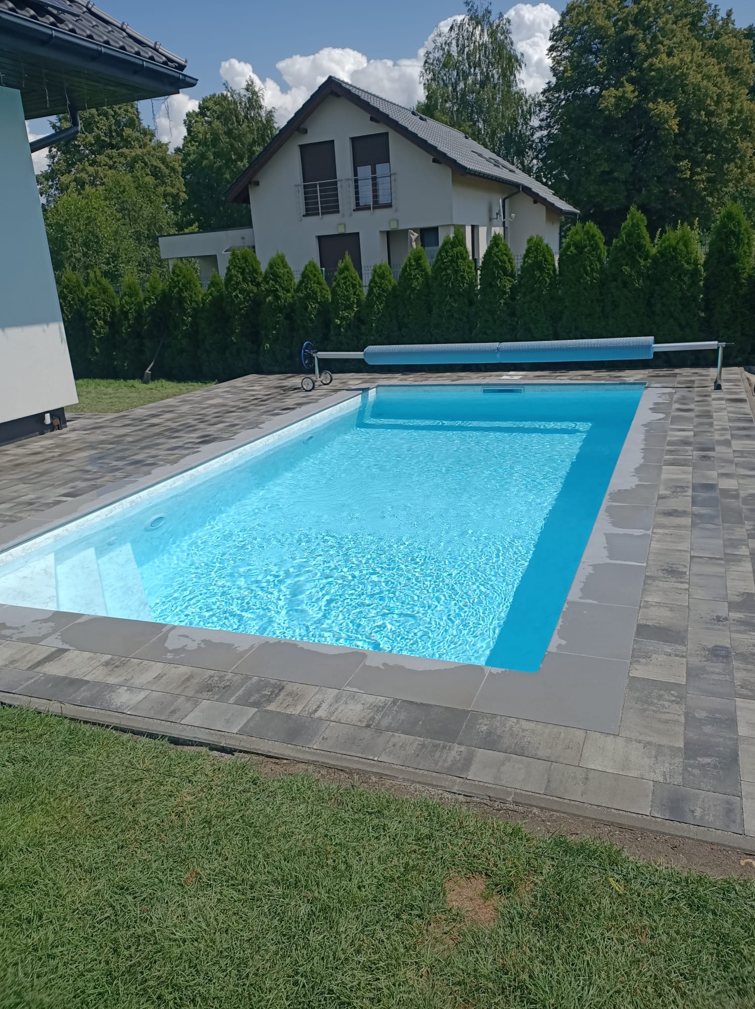 Błękitny basen z szarą kostką brukową w ogrodzie, przykryty częściowo zwiniętą roletą. W tle dom z balkonem i zielone tuje. Słoneczny dzień.