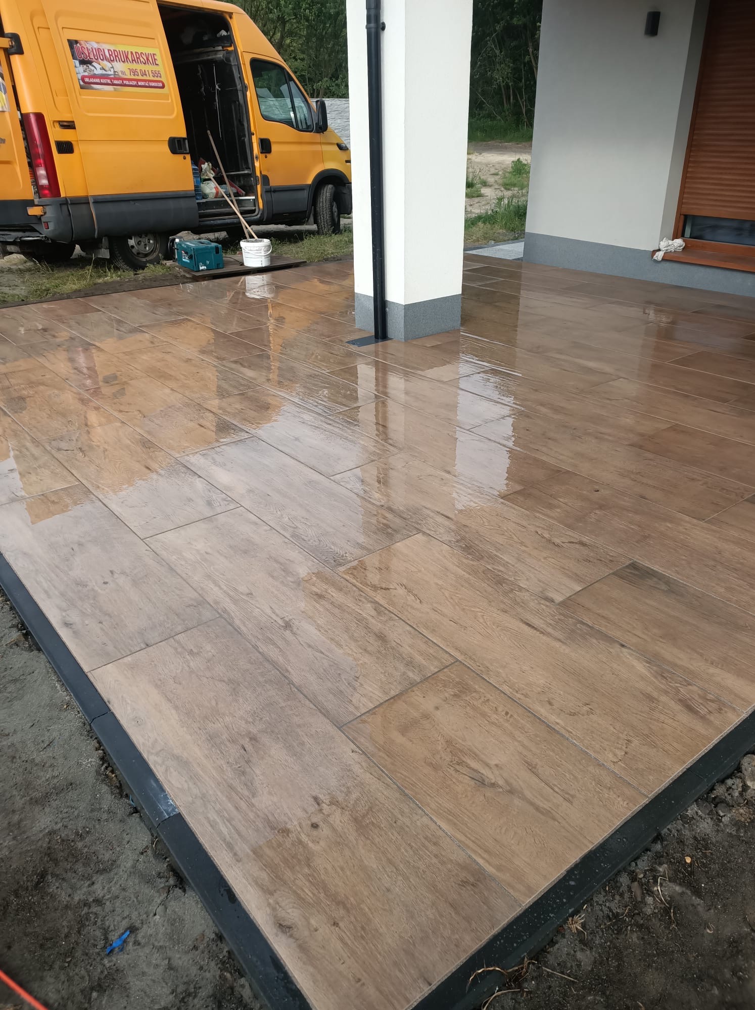 Ułożony taras z płytek imitujących drewno, obramowany czarnym obrzeżem. Na tarasie widoczne odbicia, w tle żółty samochód dostawczy z reklamą usług brukarskich.