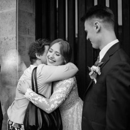 Wera Wysk Fotografia - Czarno-białe zdjęcie uśmiechniętej panny młodej w koronkowej sukni, obejmowanej przez gościa. W tle pan młody i osoba filmująca smartfonem. Ujęcie z reportażu ślubnego.