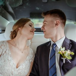 Wera Wysk Fotografia - Młoda para w samochodzie: panna młoda w koronkowej sukni spogląda z uśmiechem na pana młodego w garniturze z butonierką. Romantyczny moment w podróży.