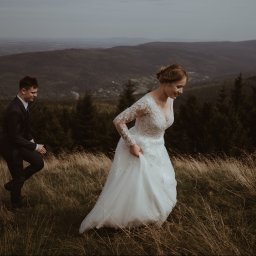 Wera Wysk Fotografia - Para młoda w plenerze górskim: panna młoda w koronkowej sukni, pan młody w garniturze, spacerują po łące z wysoką trawą, w tle zalesione wzgórza i mglista panorama.