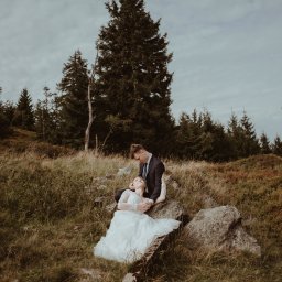 Wera Wysk Fotografia - Para młoda w plenerze górskim: panna młoda w koronkowej sukni leży na kamieniu, pan młody w garniturze obejmuje ją, w tle las iglasty i pochmurne niebo. Romantyczna sesja.