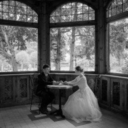 Wera Wysk Fotografia - Czarno-białe zdjęcie pary w eleganckich strojach siedzącej przy okrągłym stoliku w drewnianej altanie z dużymi oknami, przez które widać drzewa.
