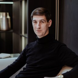 Wera Wysk Fotografia - Mężczyzna w czarnym golfie siedzi na jasnej sofie, z zegarkiem na ręku. W tle ciemne półki z dekoracjami. Portret w neutralnym oświetleniu.