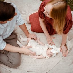 Wera Wysk Fotografia - Rodzinne ujęcie z góry: rodzice czule spoglądają na leżącą na kocu córeczkę w kremowej sukience i z kokardą. Delikatne światło podkreśla bliskość i intymność chwili.