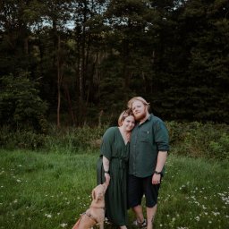 Wera Wysk Fotografia - Zakochana para w zielonych strojach pozuje z psem na łące pełnej białych kwiatów, w tle ciemny las. Romantyczna atmosfera, naturalne światło.