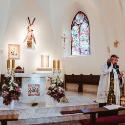 Wera Wysk Fotografia - Ksiądz z mikrofonem w dłoni podczas ceremonii w kościele. W tle ołtarz z kwiatami, witraże i ławki. Ujęcie z boku, wnętrze kościoła oświetlone naturalnym światłem.