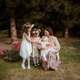 Wera Wysk Fotografia - Uśmiechnięta mama z niemowlęciem na kolanach, otoczona dwiema dziewczynkami w wiankach na głowach, w plenerze z zielenią i kwiatami. Ciepłe, naturalne światło.
