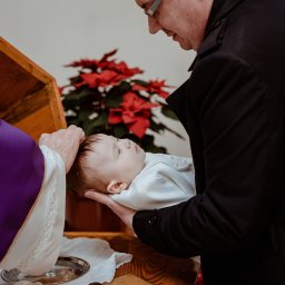 Wera Wysk Fotografia - Chrzest dziecka: ksiądz polewa wodą główkę niemowlęcia trzymanego przez ojca, w tle czerwone kwiaty i drewniana skrzynia.