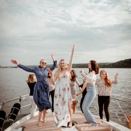 Wera Wysk Fotografia - Grupa kobiet świętuje na jachcie, w tle woda i pochmurne niebo. Uśmiechnięte, rozbawione osoby w ruchu, w letnich stylizacjach. Fotografia z perspektywy dziobu łodzi.