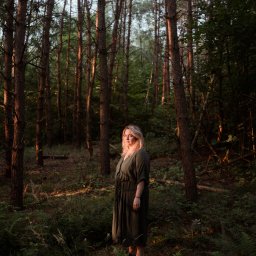 Wera Wysk Fotografia - Portret kobiety w leśnej scenerii. Blond włosy, długa sukienka w odcieniach zieleni. Drzewa tworzą naturalną ramę, a delikatne światło podkreśla atmosferę tajemniczości.