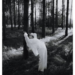 Wera Wysk Fotografia - Czarno-biała sesja kobieca w lesie: kobieta w białym kombinezonie opiera się o drzewo, z zamyślonym wyrazem twarzy, otoczona wysokimi drzewami i trawą.