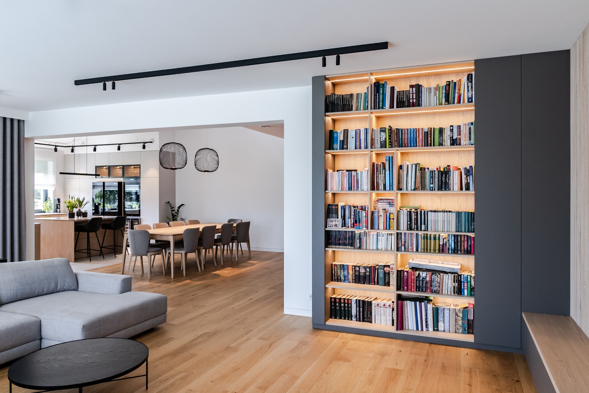 Jasne wnętrze domu z drewnianą podłogą, szarą sofą i regałem z książkami podświetlonym od wewnątrz. W tle widoczna kuchnia i jadalnia.