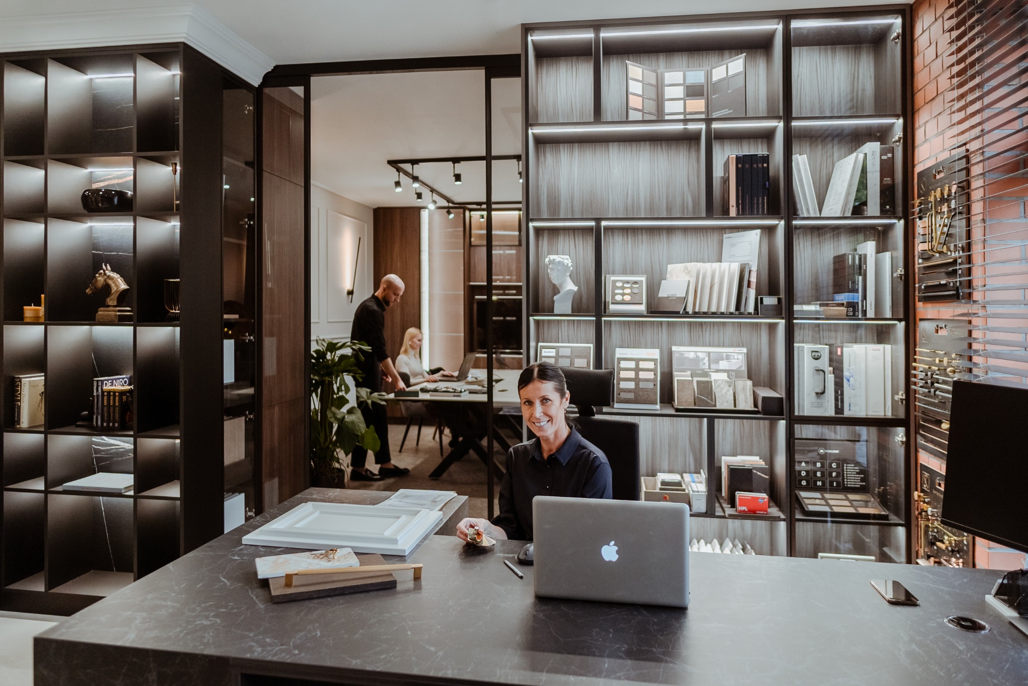 Uśmiechnięta kobieta siedzi za biurkiem z laptopem w eleganckim biurze projektowym. W tle regały z próbkami materiałów i współpracownicy przy pracy.