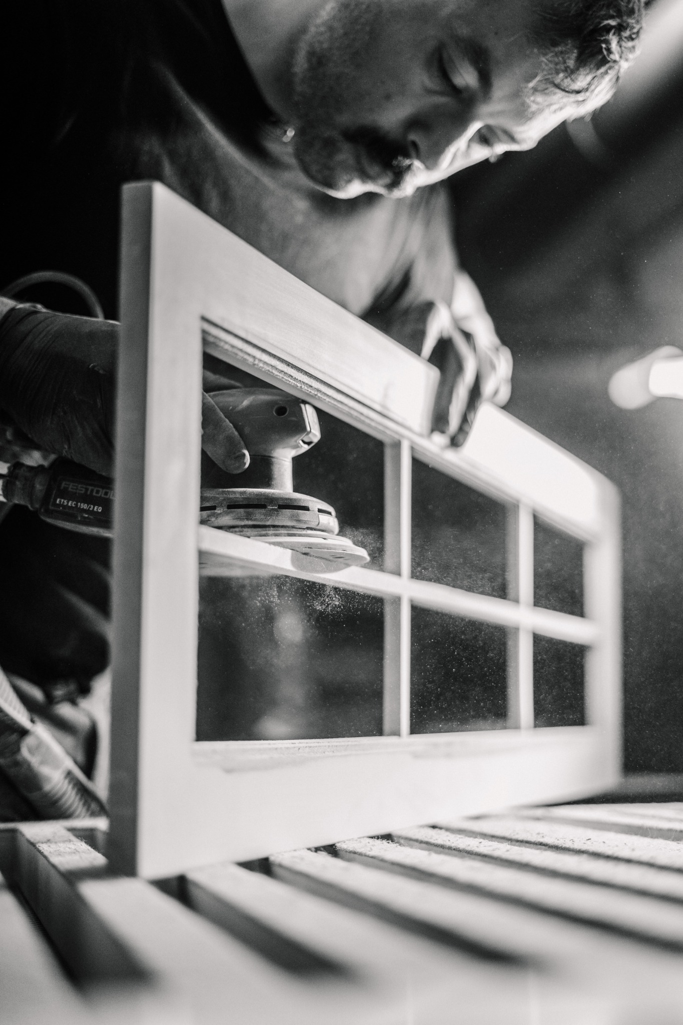 Stolarz szlifuje ramę okienną szlifierką Festool. Czarno-białe zdjęcie z pyłem unoszącym się w powietrzu, ukazujące precyzję obróbki drewna.